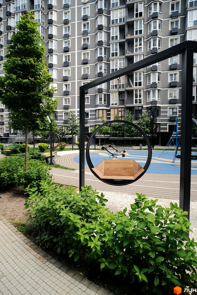 Багатоповерховий будинок Житлового комплексу Гранд Бурже з сірим фасадом. Прибудинкова територія з зеленими насадженнями та дитячим майданчиком.