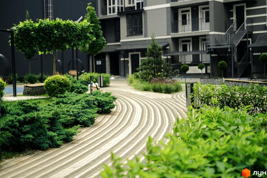 Сірий багатоквартирний будинок Житлового комплексу Гранд Бурже з балконами та зовнішніми сходами. Прибудинкова територія має доріжки та зелені насадження.