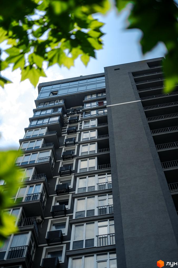 Висотна будівля Житлового комплексу Гранд Бурже з темно-сірим фасадом, численними вікнами та балконами. На передньому плані видно зелене листя дерев.