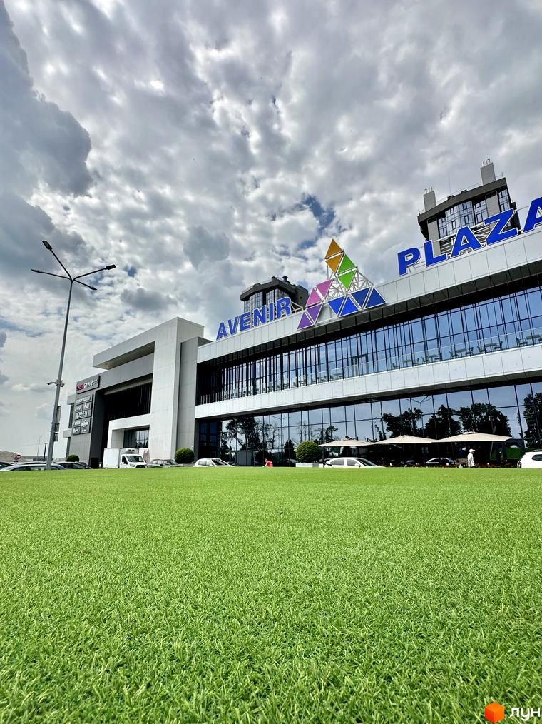 Велика будівля з дзеркальним фасадом та яскравою вивіскою, що є частиною інфраструктури Житлового комплексу Гранд Бурже. Перед нею розкинувся доглянутий зелений газон.