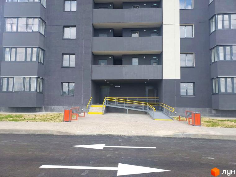 Зовнішній вигляд Житлового комплексу Оазис: темно-сірий фасад, засклені балкони, вхід з пандусом та лавками.