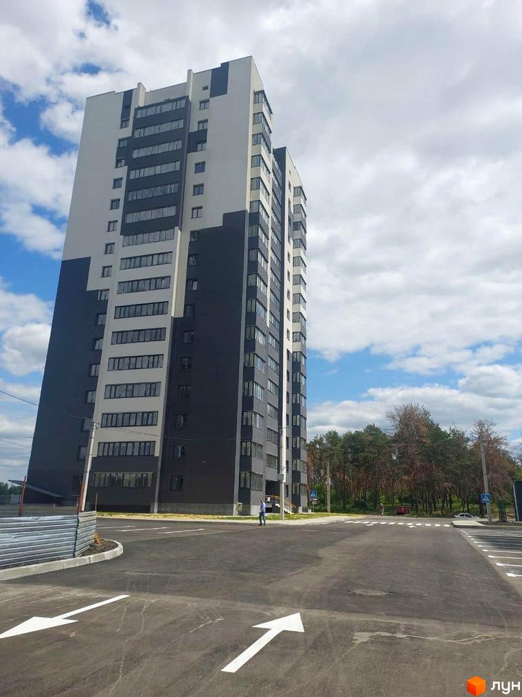 Багатоповерховий будинок Житлового комплексу Оазис у сіро-білих тонах. Перед будинком асфальтована територія з розміткою та дерева на задньому плані.