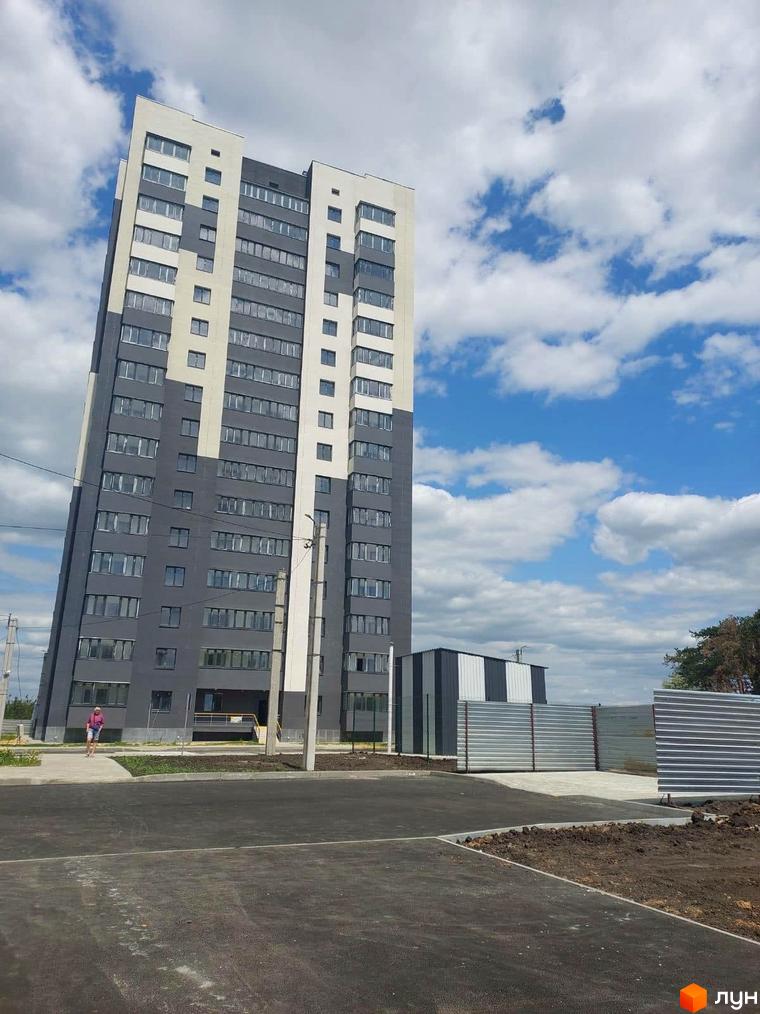 Новобудова Оазис: багатоповерховий будинок сірого кольору з бежевими вставками. Перед ним асфальтована дорога та частково облаштована територія.