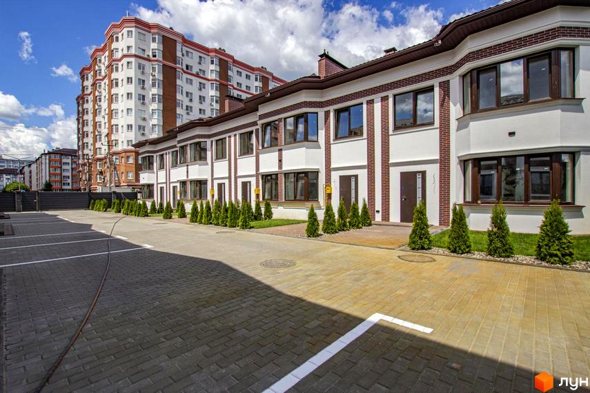 Таунхауси на Львівському - це двоповерхові будинки з білою та цегляною обробкою, розташовані на брукованій прибудинковій території з зеленими насадженнями. На задньому плані видно багатоповерховий житловий будинок.