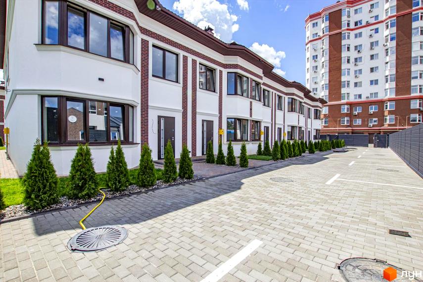 Таунхауси на Львівському: фасади з білою штукатуркою та червоною цеглою, вікна з темними рамами, зелені туї та парковка перед будинками.