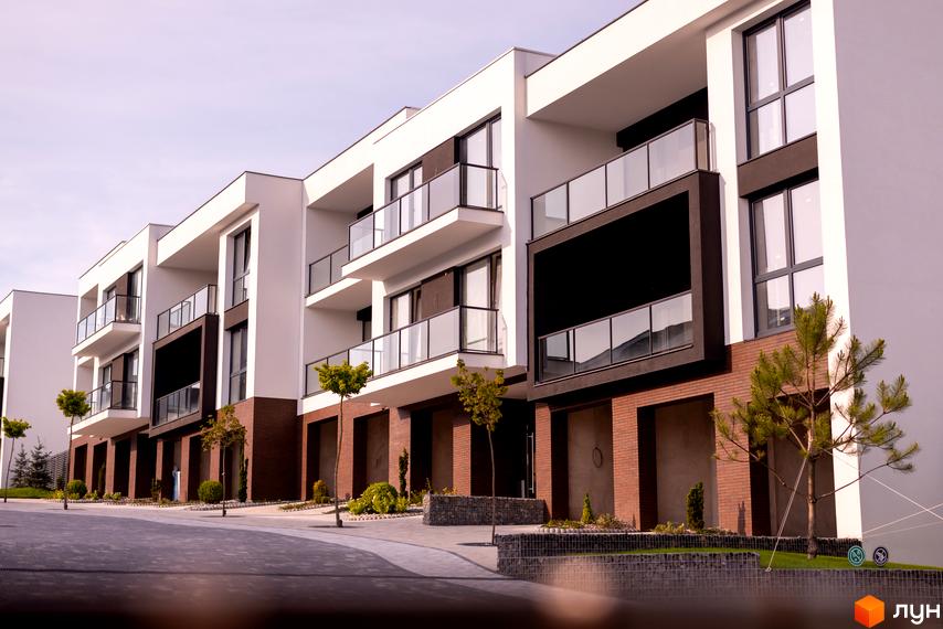 Зовнішній вигляд житлового комплексу SHERWOOD з білими фасадами, темними акцентами та скляними балконами. Навколо новобудови доглянута прибудинкова територія з деревами та кущами.