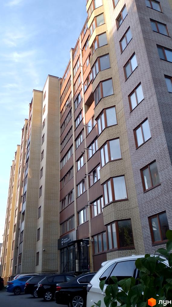 Зовнішній вигляд багатоповерхового будинку з цегляним фасадом та балконами. На передньому плані припарковані автомобілі.