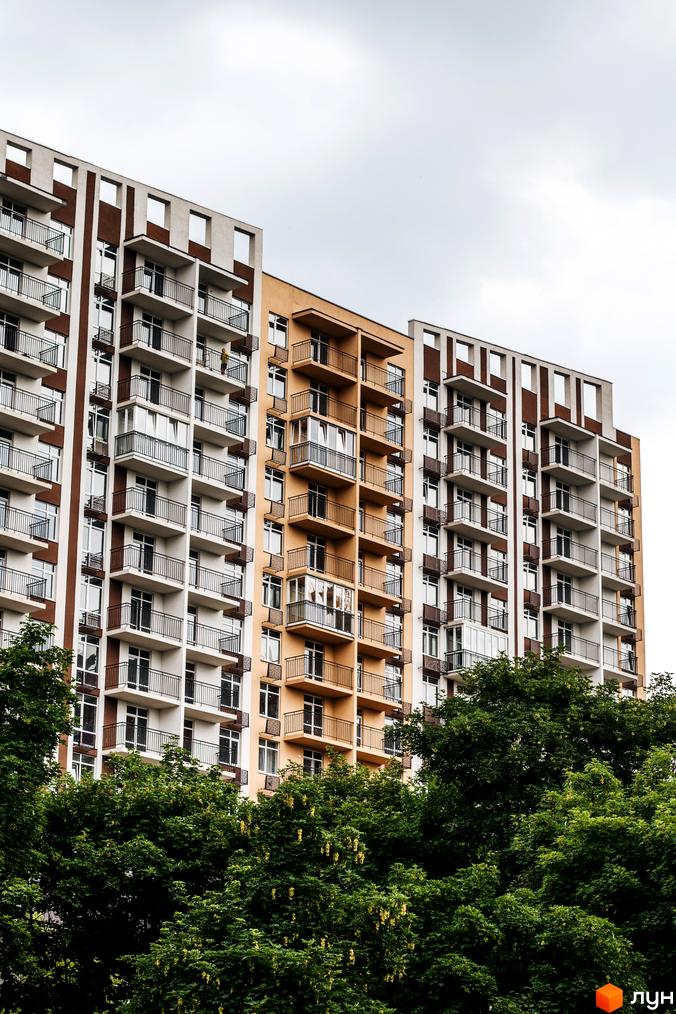 Новобудова ЖК Кришталеві джерела з балконами та вікнами на тлі хмарного неба. Зелені дерева перед фасадом будівлі.