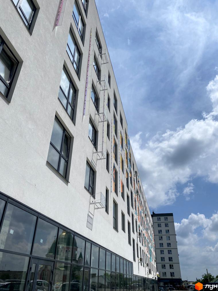Висотна будівля ЖК Синергія Glass з великими вікнами та балконами. На фасаді кольорові акценти, навколо будівлі видно хмари та небо.