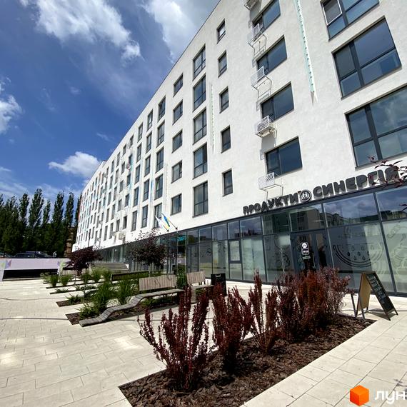 Зовнішній вигляд будівлі житлового комплексу Синергія Glass з великими вікнами та скляними вхідними групами. На передньому плані доглянута прибудинкова територія з рослинами та лавками.