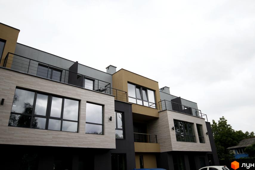 Фасад Таунхаусів Rusaniv Gardens з багатоповерховим дизайном, світлими та темними панелями, великими вікнами та балконами. Поруч видно зелені насадження.