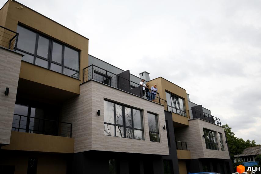 Зовнішній вигляд таунхаусів Rusaniv Gardens з великими вікнами та балконами. На балконі стоять люди.