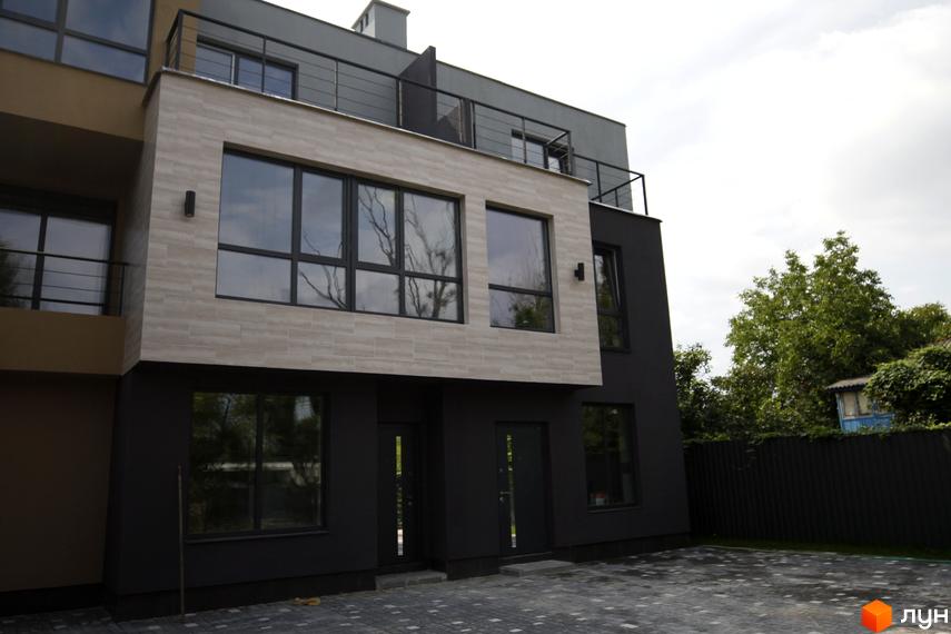 Зовнішній вигляд таунхаусів Rusaniv Gardens з великими вікнами та темними фасадами. На передньому плані - бруківка.