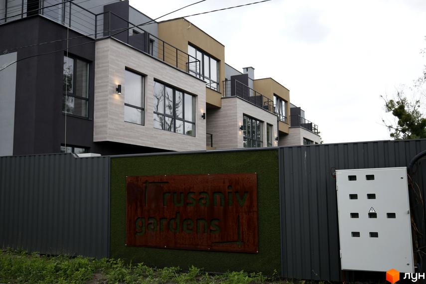 Зображення таунхаусів Rusaniv Gardens з сіро-бежевими фасадами та великими вікнами. Перед ними огорожа з вивіскою проєкту та зеленою панеллю.