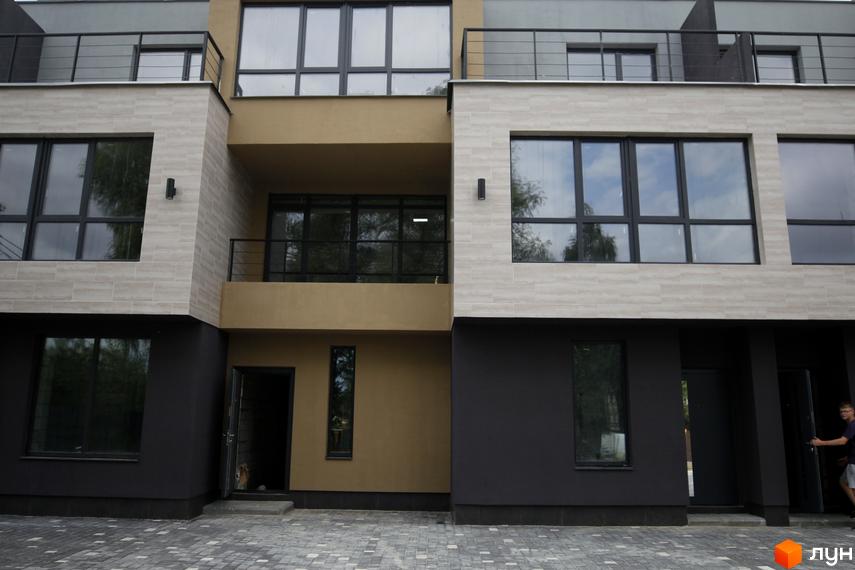 Фасад таунхаусів Rusaniv Gardens з темно-сірими та бежевими елементами, великими вікнами та вимощеною бруківкою прибудинковою територією.