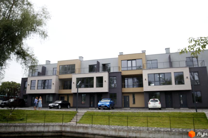 Таунхауси Rusaniv Gardens з багатоповерховими фасадами світлих та темних кольорів, великими вікнами. Перед ними зелений газон, доріжка та водойма.