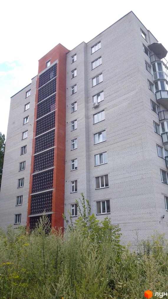 Зовнішній вигляд багатоповерхового будинку з цегляним оздобленням та скляними елементами. На передньому плані видно густі зелені насадження.