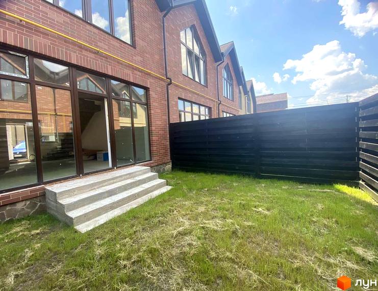 Цегляний будинок з панорамними вікнами та терасою. На передньому плані газон та сходи, за будинком чорний паркан.