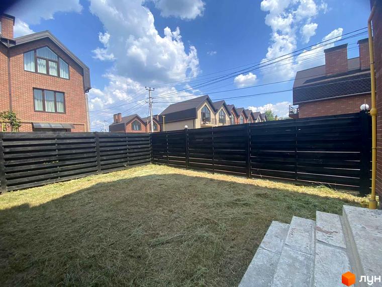 Цегляний будинок з панорамними вікнами та темним парканом. На прибудинковій території газон з сухою травою.