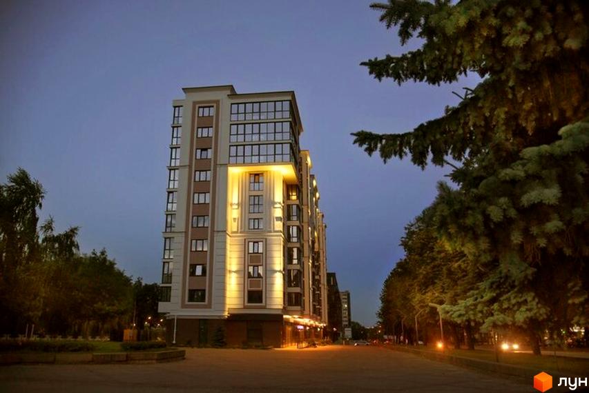 Житловий комплекс GREAT HOUSE з підсвіткою фасаду та панорамними вікнами на тлі вечірнього неба. Навколо комплексу зелені насадження та освітлена вулиця.