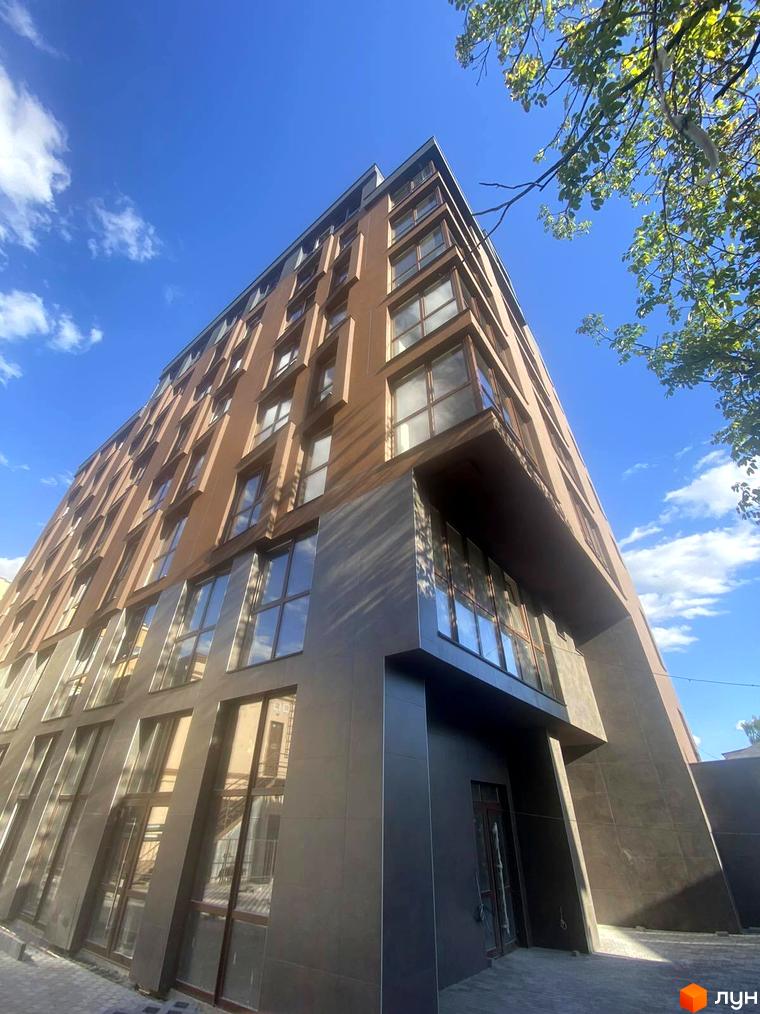 Фасад житлового комплексу Elegant з численними вікнами та коричневою цегляною обробкою, на тлі блакитного неба з хмарами. Територія навколо будинку вимощена плиткою.