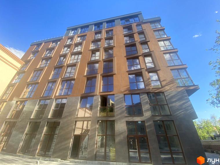 Фасад житлового комплексу Elegant з коричневою та сірою облицовкою, великими вікнами, що відбивають небо та хмари. Будинок має багатоповерхову структуру під блакитним небом.