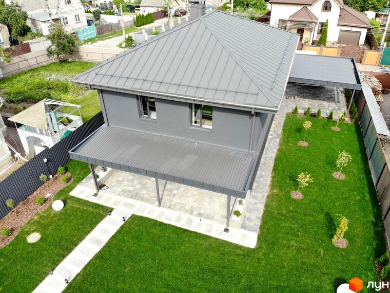 Двоповерховий будинок сірого кольору з металевим дахом та критою терасою. Прибудинкова територія Котеджного містечка Kulbaba Petropavlivka має доглянутий газон та вимощені доріжки.