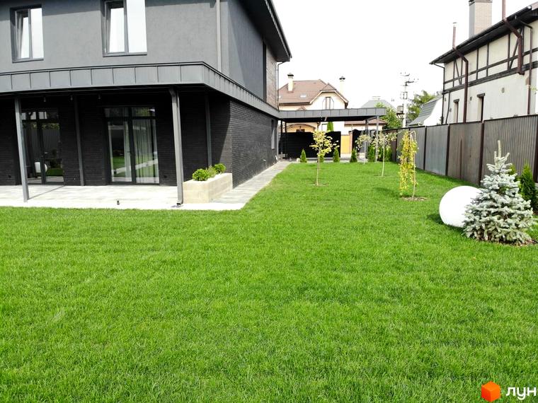 Фасад будинку Котеджного містечка Kulbaba Petropavlivka з темною обробкою та великим газоном. На території ростуть молоді дерева та ялинка.