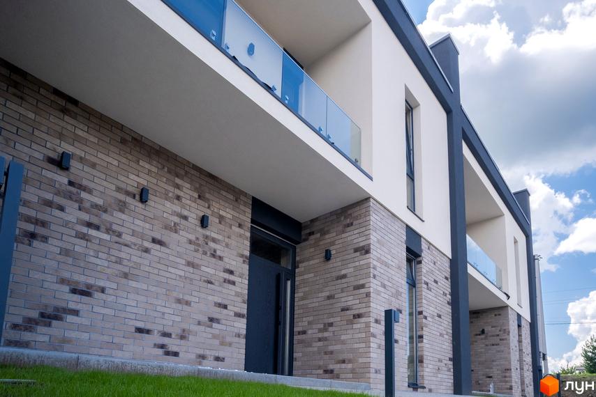 Фасад будинку в Котеджному містечку Квадрат TOWN з цегляною кладкою та скляними балконами, зелена трава на прибудинковій території, блакитне небо з хмарами.