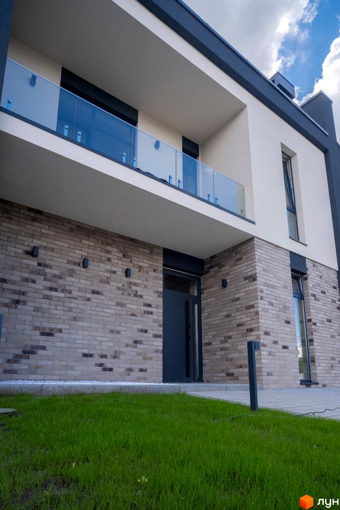 Зображення будинку в Котеджному містечку Квадрат TOWN. Фасад з цегли, балкон з прозорим склом та зелений газон перед будинком.