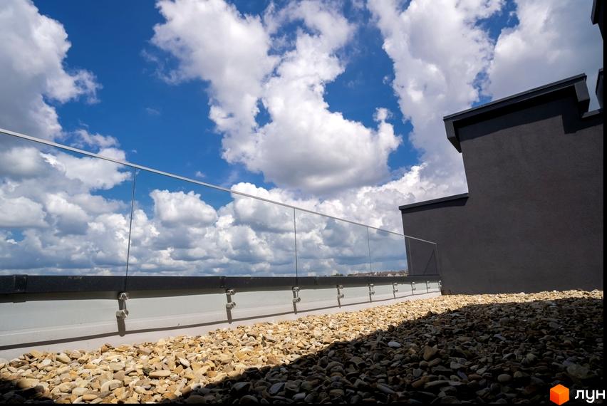 Котеджне містечко Квадрат TOWN: фрагмент даху будинку з темно-сірими стінами та скляним огородженням, навколо гравійна поверхня.