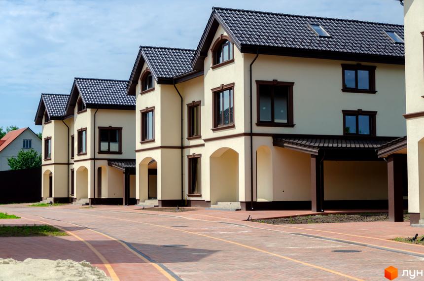 Котеджне містечко КМ Vita Energy residence - фасади двоповерхових будинків світло-бежевого кольору з темними дахами та вікнами. Поруч брукована дорога та зелені насадження.