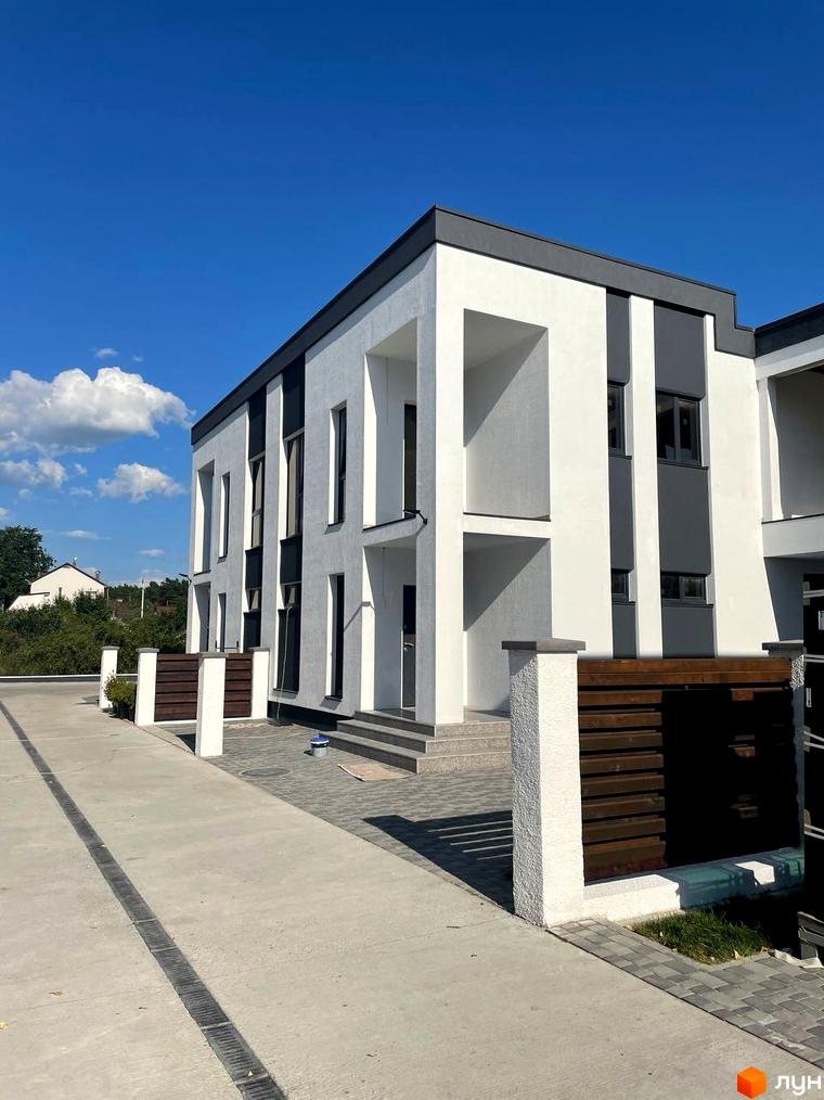 Котеджне містечко КМ Перлина Процева: фасади будинків з білою та сірою обробкою, великі вікна, прибудинкова територія з бруківкою та дерев'яними парканами.