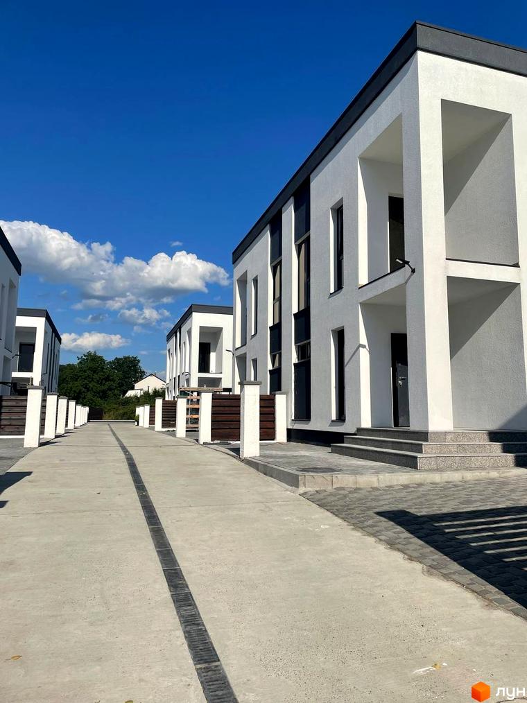 Котеджне містечко Перлина Процева: фасади білих будинків з чорними елементами та бетонна алея з ліхтарями. Навколо зелені насадження та блакитне небо з хмарами.