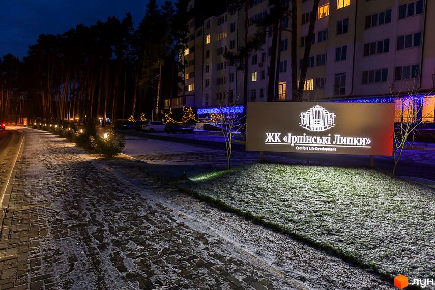 Житловий комплекс Ірпінські Липки вночі. Зовнішній вигляд будівлі з підсвіченими вікнами та вивіскою. Прибудинкова територія з освітленням та снігом на бруківці.