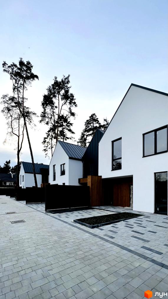 Котеджне містечко КМ Toscana House 2 з білими фасадами та темними дахами, розташоване біля соснового лісу, з вимощеною сірою плиткою прибудинковою територією.