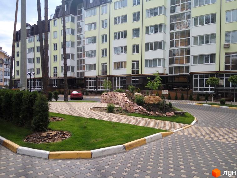 Житловий комплекс Ірпінські Липки з жовто-білим фасадом та прибудинковою територією з бруківки та зелених насаджень. Автомобіль червоного кольору припаркований біля будинку.