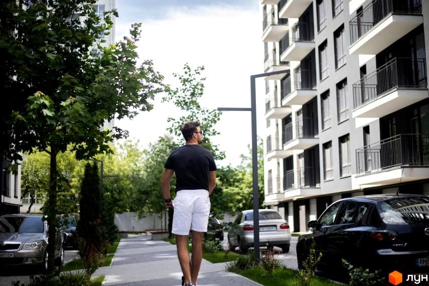 Житловий комплекс Comfort Park. Чоловік на самокаті їде вулицею повз сучасну новобудову з балконами та прибудинковою територією з деревами та автомобілями.