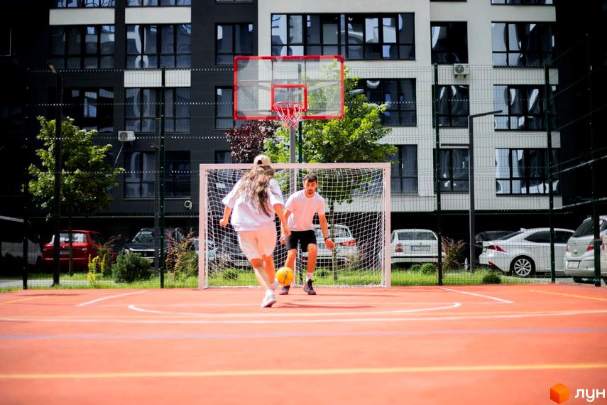 Житловий комплекс Comfort Park. На території комплексу розташований спортивний майданчик, де молода пара грає у футбол. На задньому плані видно сучасні багатоквартирні будинки та припарковані автомобілі.