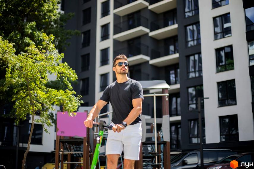 Житловий комплекс Comfort Park: чоловік на самокаті на фоні багатоповерхових будинків з балконами та вікнами. Прибудинкова територія з деревами та дитячим майданчиком.