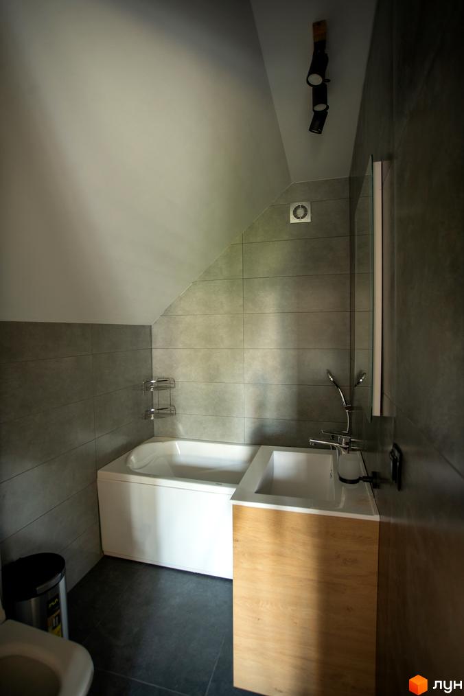 Ванна кімната в будинку КМ Gutenberg з білою ванною та душем, сірою плиткою та дерев'яною тумбою під раковину.