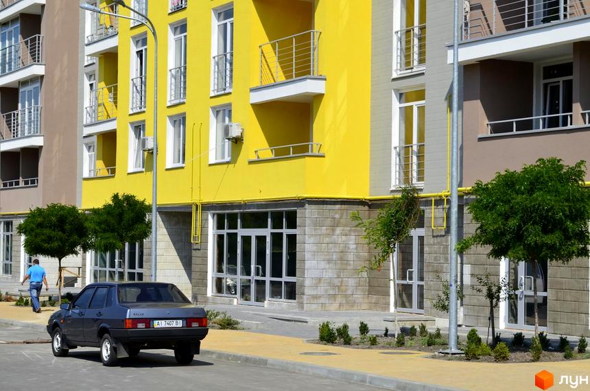 Житловий комплекс Столичний. Фасад жовтої новобудови з балконами та кондиціонерами. Поруч прибудинкова територія з деревами та припаркованим автомобілем.