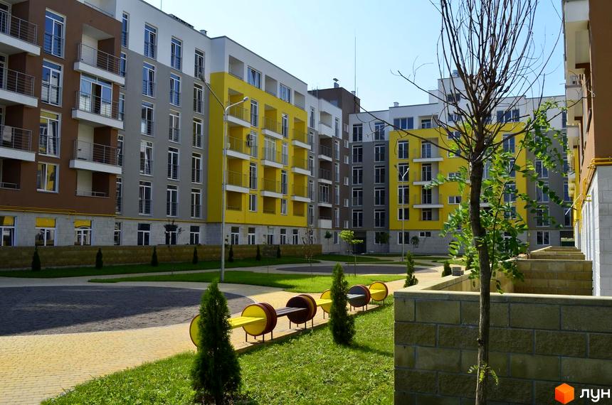 Житловий комплекс Столичний: багатоповерхові будинки з жовтими та сірими фасадами, балконами та великими вікнами. Прибудинкова територія з лавочками, деревами та газоном.