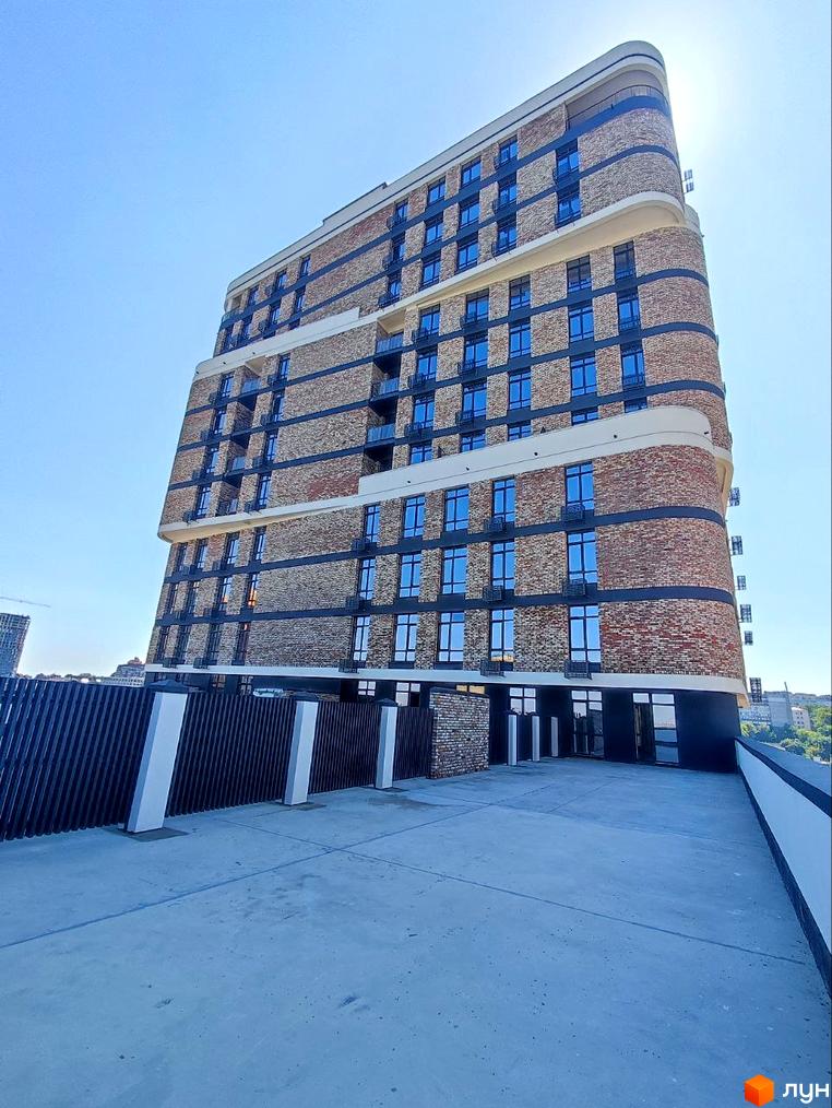 Новобудова Loft White з цегляним фасадом, округлими кутами та синіми вікнами. Перед будинком бетонний майданчик та темний паркан.