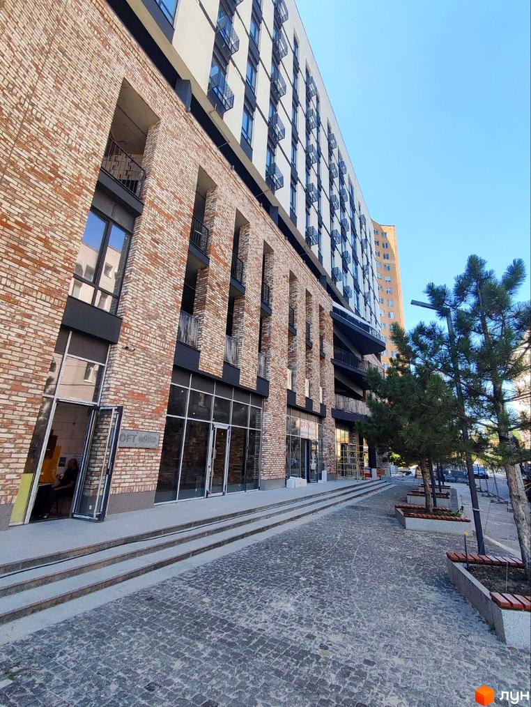 Зовнішній вигляд житлового комплексу Loft White з цегляним фасадом та великими вікнами. Поруч розташована брукована алея з деревами та лавками.