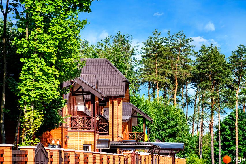 Котеджне містечко КМ Парк хаус. Цегляний будинок з коричневим дахом та дерев'яними балконами, оточений зеленню та соснами.