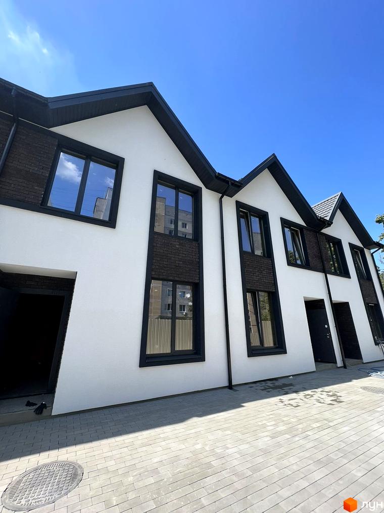 Білосніжні таунхауси з чорними вікнами та дахом на тлі блакитного неба. Бруківка на прибудинковій території.