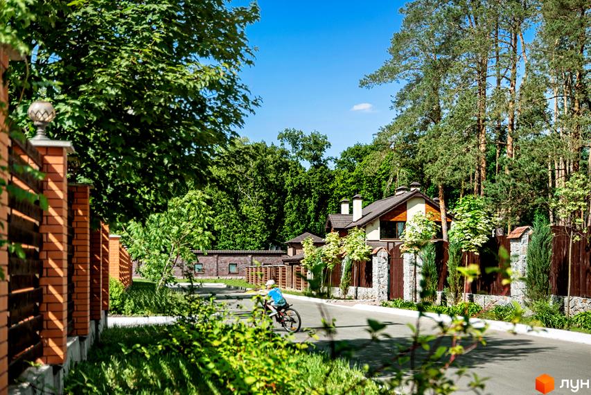Котеджне містечко Парк Хаус: двоповерховий будинок з коричневим дахом та білими стінами, оточений деревами та парканом. Дитина їде на велосипеді по асфальтованій дорозі.