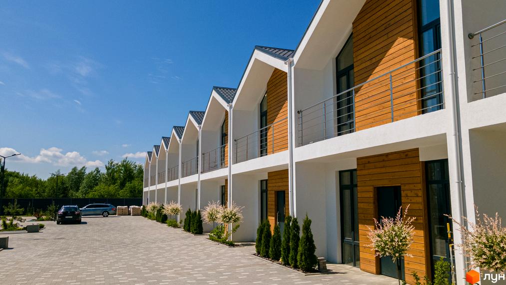 Котеджне містечко КМ Prydesennya Eco Village: фасади будинків з дерев'яними елементами та балконами, доглянута прибудинкова територія з парковкою та зеленими насадженнями.