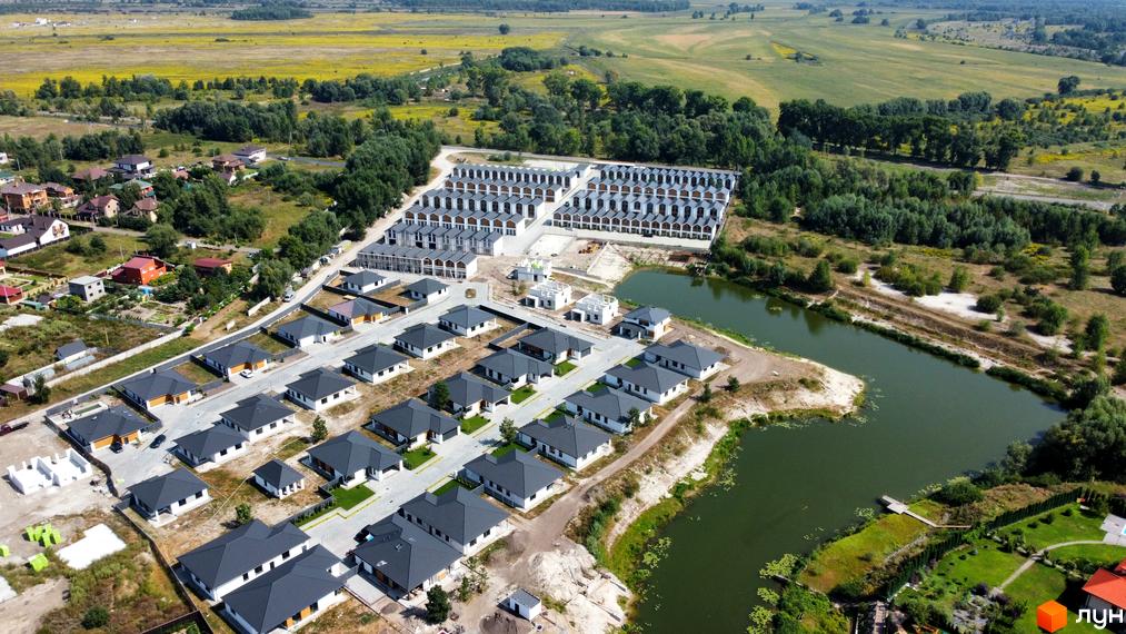 Вигляд з висоти на Котеджне містечко Prydesennya Eco Village з будинками з темними дахами та озером поруч. Навколо комплексу зелені поля та ліс.